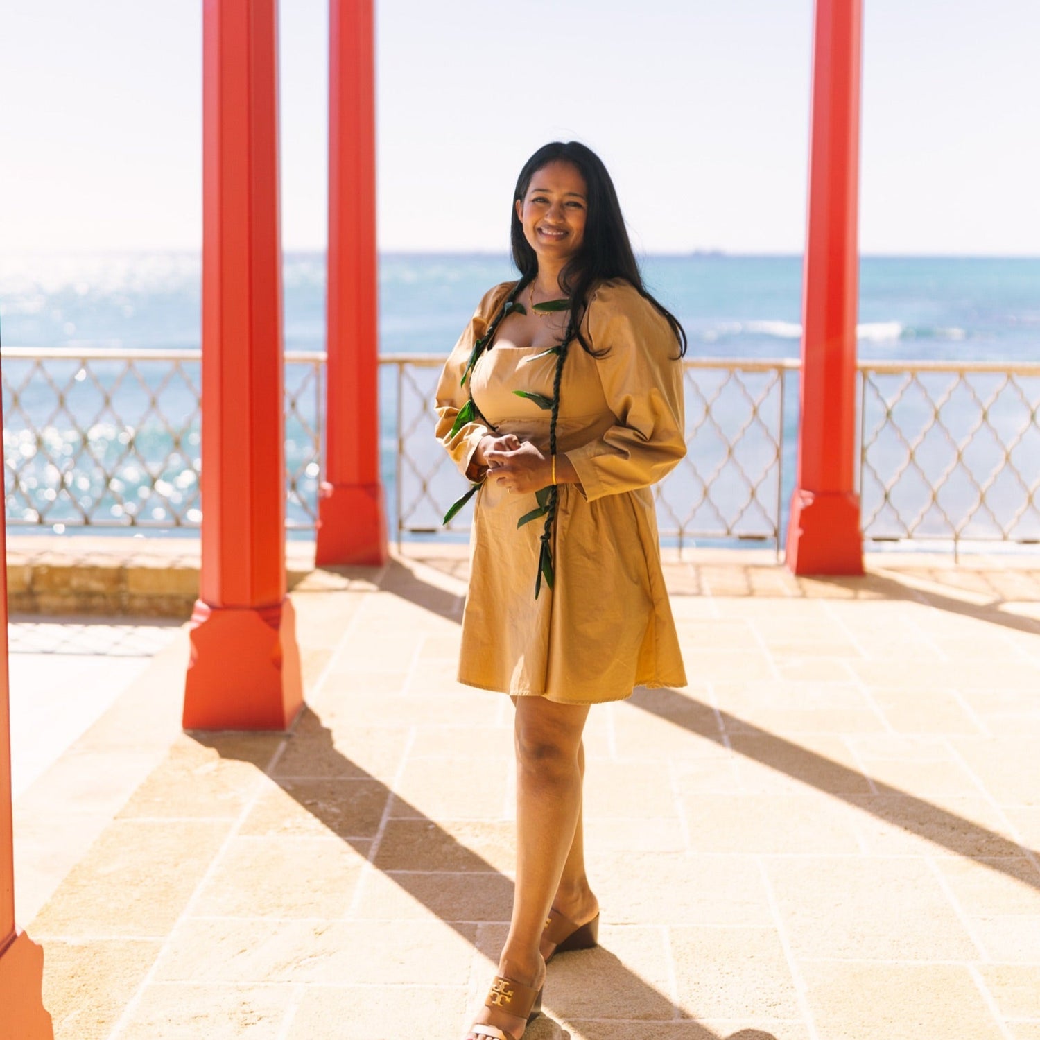 Dr. Suma in a yellow dress standing on a balcony with red columns and ocean view on oahu hawaii