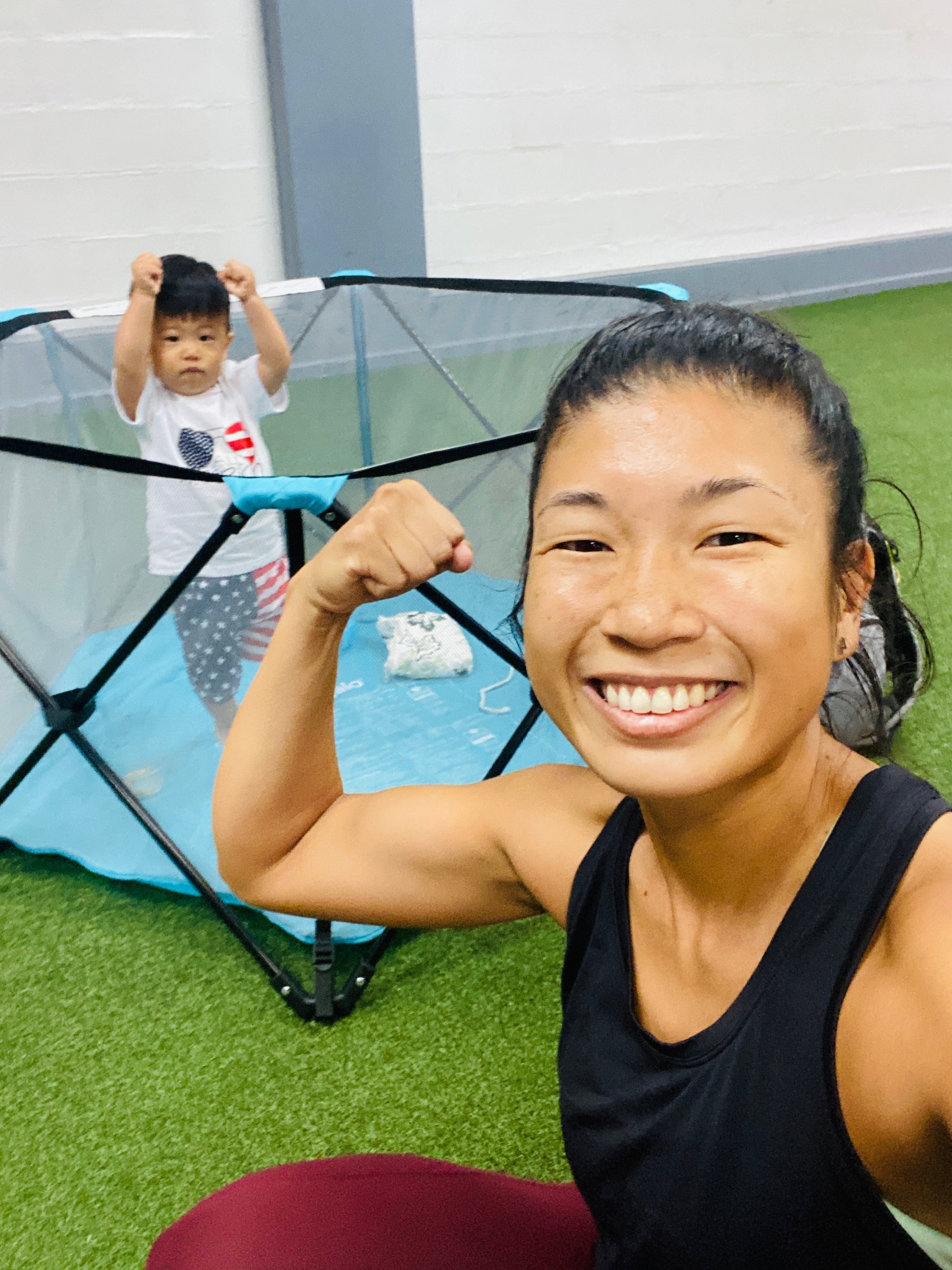 mom flexing her arm muscles in front of a child playing inside a playpen at the gym.