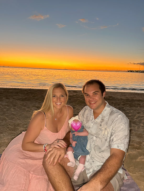 Couple sitting on a hawaii beach at sunset holding a baby girl with a pink heart covering her face.