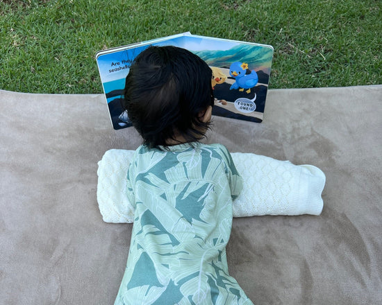 baby in a green patterned onesie lying on a blanket doing tummy time and reading a book outdoors. 