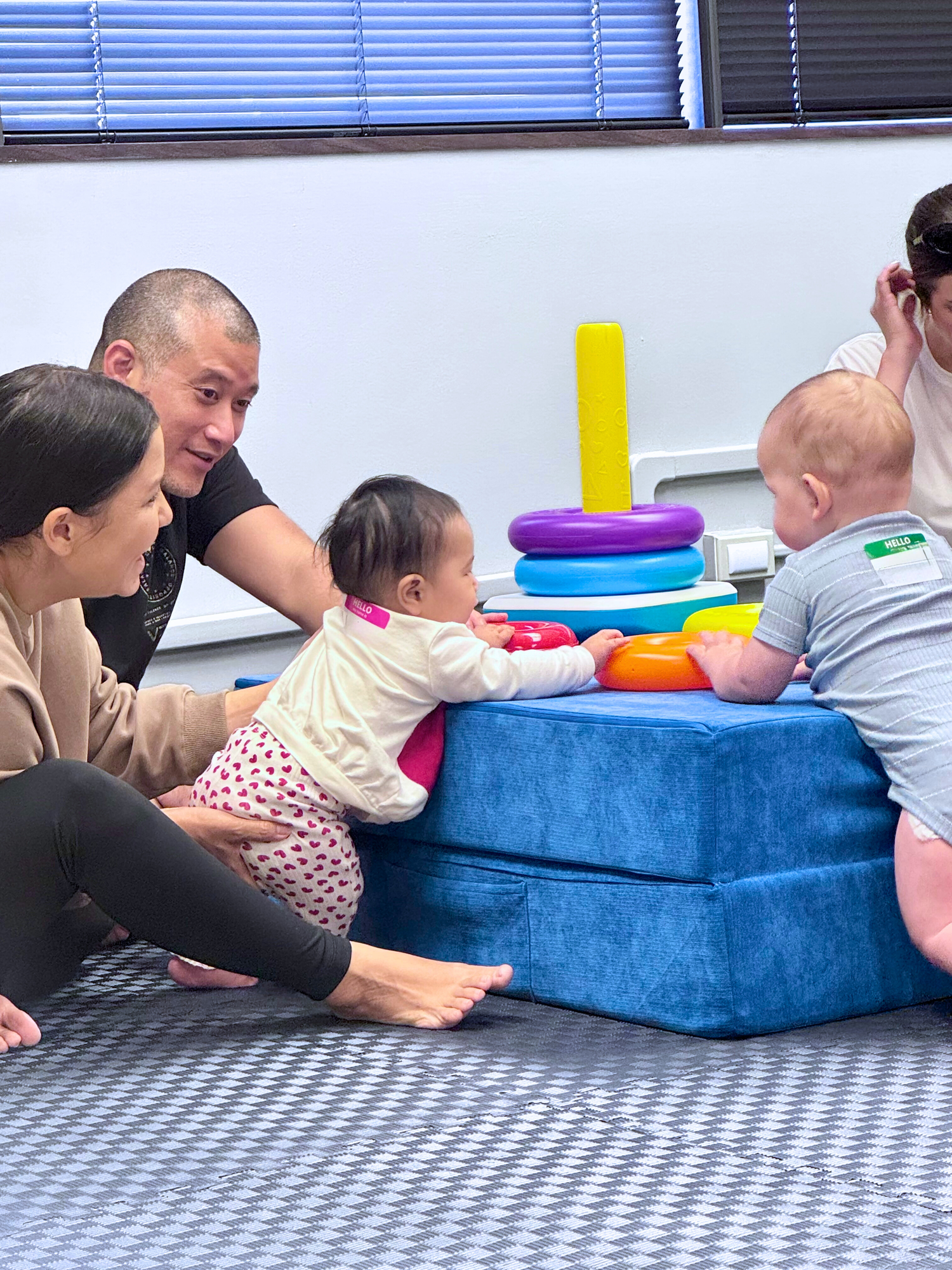 Family with two babies playing with colorful toys on a blue mat.